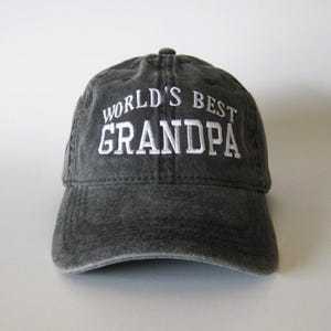 以下が含まれることがあります： 白の「WORLD'S BEST GRANDPA」の刺繍が施された濃いグレーの野球帽。帽子はカーブしたつばと布製のストラップ留めが付いています。祖父母向けのカジュアルなアクセサリー。