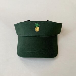 May include: A dark green visor with a curved brim and a small, embroidered pineapple in yellow and green on the front. The visor is made of a sturdy fabric and has a black strap.
