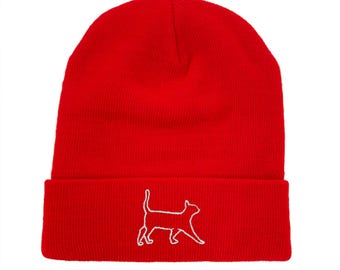 Puede incluir: Un gorro rojo brillante con un borde doblado. El gorro presenta un contorno blanco bordado de un gato caminando por el borde. El gorro está hecho de un material de punto suave.