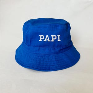 Może przedstawiać: Królewski niebieski kapelusz typu bucket z białym haftem "PAPI". Kapelusz wykonany jest z miękkiego materiału i ma klasyczny, swobodny styl. Idealny do ochrony przed słońcem lub jako element mody.