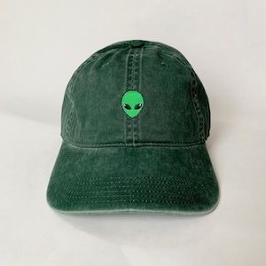 Puede incluir: Una gorra de béisbol verde oscuro con una cara de extraterrestre verde bordada en la parte delantera.