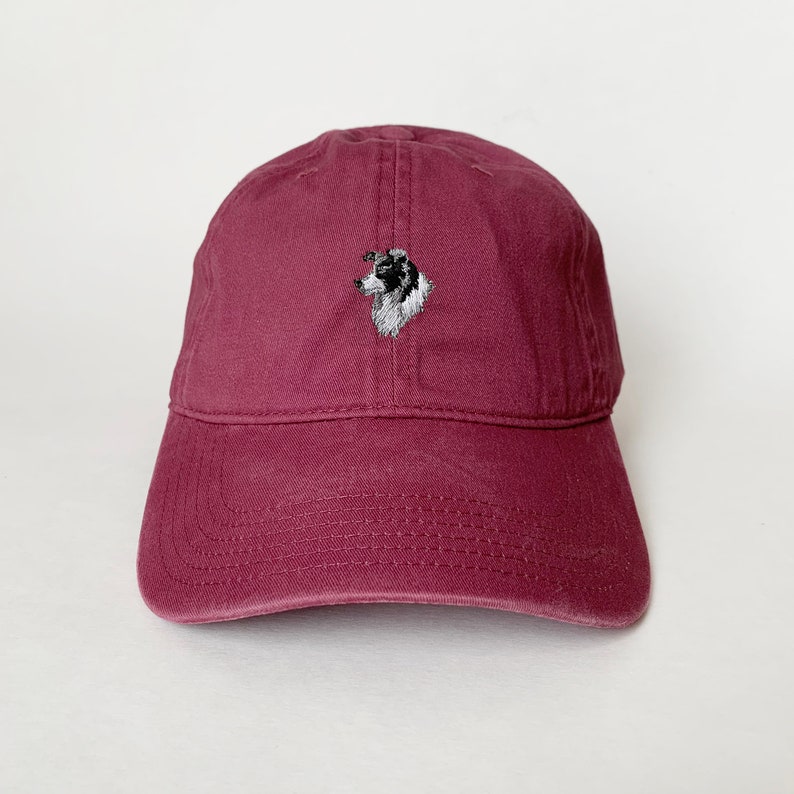 Puede incluir: Una gorra de b&eacute;isbol de color burdeos con un border collie blanco y negro bordado en la parte delantera.