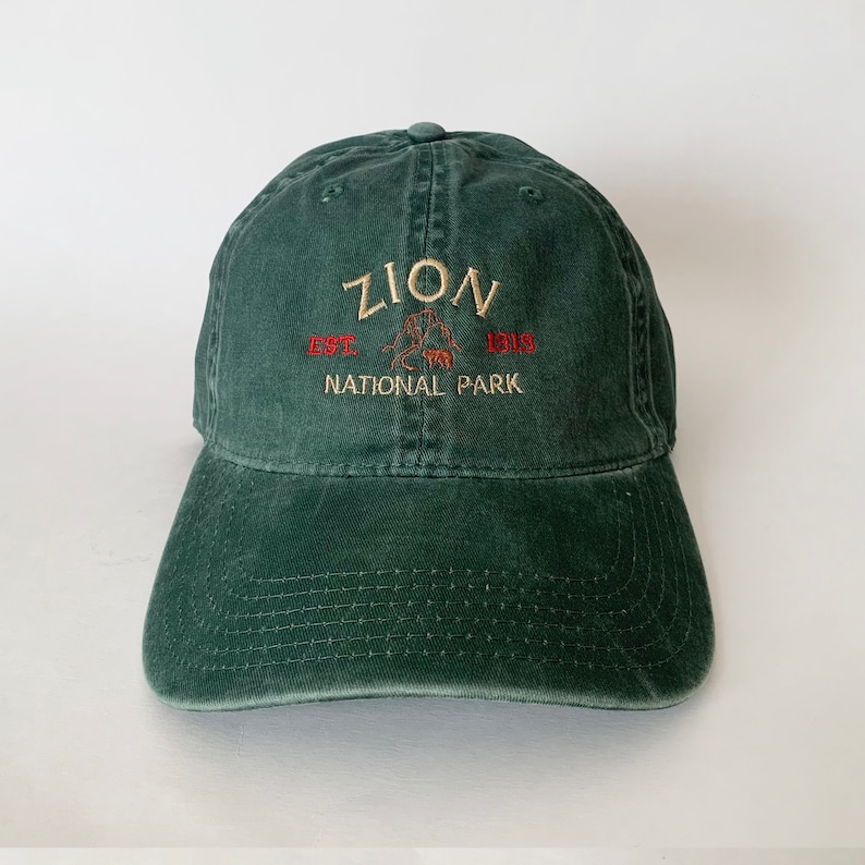 May include: A dark green baseball cap with an embroidered logo that reads "Zion Est. 1919 National Park".