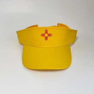 May include: A bright yellow visor with a curved brim and a red emblem. The emblem is a stylized cross with four arms, each ending in a straight line. The visor is made of a sturdy fabric.