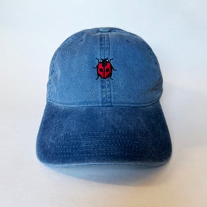 May include: A blue baseball cap with a curved peak. The cap has a red and black embroidered ladybird design on the front. The hat is made of a soft, washed fabric.