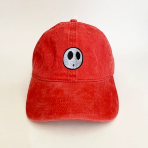 Puede incluir: Una gorra de béisbol roja con una cara de fantasma bordada en blanco y gris.