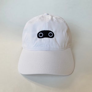 Könnte beinhalten: Eine weiße Baseballkappe mit einem schwarzen gestickten Design aus zwei Augen auf der Vorderseite.