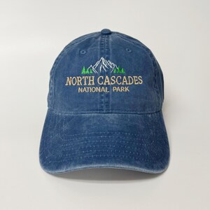 May include: A blue baseball cap with the text "North Cascades National Park" embroidered in green and tan. The design features a mountain range and trees. The cap is made of a soft, worn material.