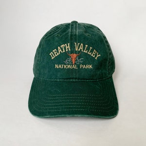 Puede incluir: Una gorra de béisbol verde oscuro con un logotipo bordado que dice "Death Valley National Park" con un diseño estilizado de cráneo y cuernos.