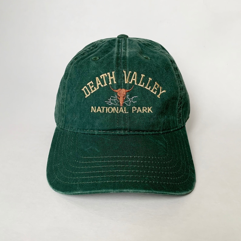 May include: A dark green baseball cap with an embroidered logo that says "Death Valley National Park" with a stylised skull and horns design.