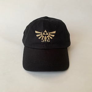 Op de afbeelding: Een zwarte baseballpet met een gouden geborduurd Triforce-symbool uit de videogame-serie The Legend of Zelda.