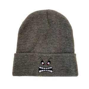 May include: Gray knitted beanie with a white embroidered angry face with red eyes.