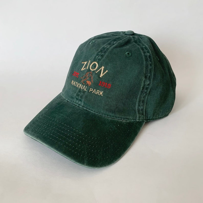 May include: A dark green cotton baseball cap with an embroidered logo that reads "ZION EST. 1919 NATIONAL PARK".