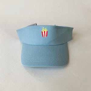 May include: A light blue visor featuring a small, centered embroidered graphic of a red and white striped popcorn bucket with yellow popcorn kernels popping out of the top. The visor has a structured front panel and a curved brim.