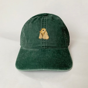 May include: A dark green baseball cap with a curved peak. The cap has an embroidered image of a Cocker Spaniel dog in tan and brown. The cap is made of a soft, worn fabric.
