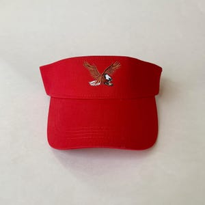 May include: A red visor with an embroidered eagle design. The visor features a curved brim and a fabric band. The eagle is depicted with outstretched wings in shades of brown, white, and black.