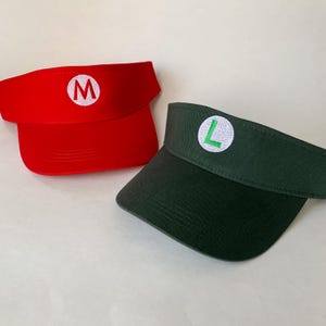 May include: A bright red visor with a white circular patch featuring a red letter M, and a dark green visor with a white circular patch featuring a green letter L. Both visors have a curved brim and an open top design for headwear.