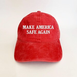 May include: Red baseball cap with the text "MAKE AMERICA SAFE AGAIN" embroidered in white. The cap has a curved brim and a six-panel design. The hat is made of a soft, washed cotton material.