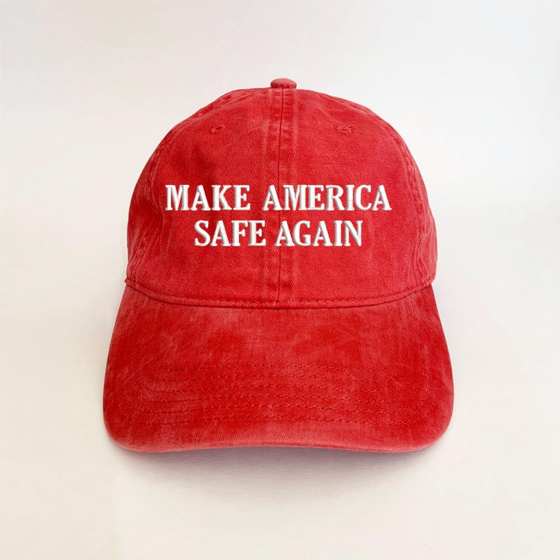 May include: Red baseball cap with the text "MAKE AMERICA SAFE AGAIN" embroidered in white. The cap has a curved brim and a six-panel design. The hat is made of a soft, washed cotton material.