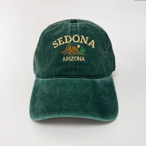 Könnte beinhalten: Dunkelgrüne Baseballkappe mit dem Wort "SEDONA" in Goldfaden über einer Wüstenlandschaft und dem Wort "ARIZONA" gestickt. Die Kappe hat einen gebogenen Schirm und einen Stoffriemenverschluss.