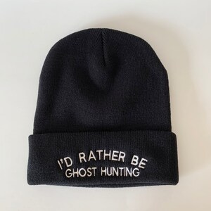 Peut inclure: Un bonnet noir avec du texte brodé blanc qui dit "J'aimerais mieux être à la chasse aux fantômes."