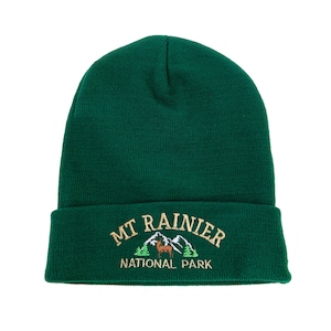 以下が含まれることがあります： 緑色のニットビーニー。ゴールドの刺繍で「MT RAINIER NATIONAL PARK」と、その下に山脈と鹿が描かれている。