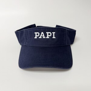 May include: Navy blue visor with the word "PAPI" embroidered in white block letters across the front. The visor has a curved brim and an adjustable strap. The visor is made of a durable fabric.