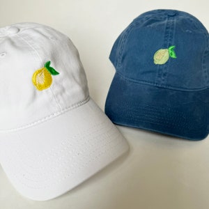Puede incluir: Dos gorras de béisbol, una blanca y otra azul, ambas con diseños de limón bordados.