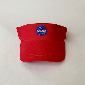Pode incluir: Viseira vermelha com o logotipo da NASA bordado na frente. O logotipo é um círculo azul com texto branco e uma faixa vermelha. A viseira é feita de tecido vermelho e tem uma aba curva.