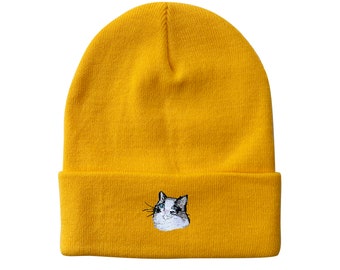 Puede incluir: Gorro de color amarillo mostaza con borde doblado. El gorro presenta un rostro de gato bordado en la parte delantera. El gato tiene pelaje blanco, marcas negras y ojos azules. El gorro está hecho de un material de punto suave.
