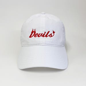 Puede incluir: Una gorra de béisbol blanca con la palabra Devils bordada en hilo rojo en la parte delantera. La letra D está estilizada con pequeños cuernos y la letra s termina en una cola puntiaguda. La gorra tiene una visera curva y una corona estructurada.