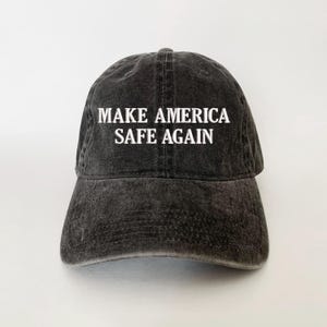 May include: A black, distressed baseball cap with the white text "MAKE AMERICA SAFE AGAIN" across the front. The cap has a curved brim and a classic six-panel design, suitable for casual wear.