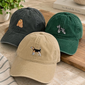 Puede incluir: Tres gorras de béisbol en gris carbón, verde bosque y color canela. Cada gorra presenta un pequeño y detallado diseño de perro bordado en el panel frontal. La gorra gris muestra un perro dorado, la verde un perro blanco y negro, y la canela un beagle.