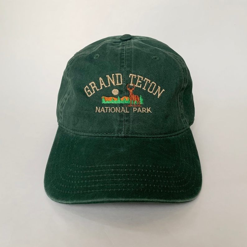 May include: A dark green baseball cap with an embroidered logo that reads "Grand Teton National Park". The logo features a silhouette of a mountain range with a deer standing in front of it.