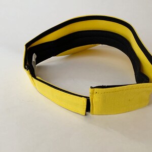 Op de afbeelding: Een gele visor met een zwarte rand en een zwarte verstelbare band. De visor is gemaakt van een zachte, comfortabele stof.
