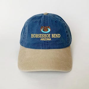 Op de afbeelding: Een marineblauwe en beige baseballpet met de tekst "HORSESHOE BEND ARIZONA" geborduurd op de voorkant. De pet heeft een hoefijzerontwerp boven de tekst. De pet heeft een gebogen klep en een knoop aan de bovenkant.