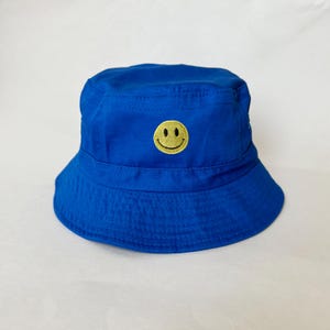 Puede incluir: Un gorro de pescador azul con una carita sonriente amarilla bordada en la parte delantera.