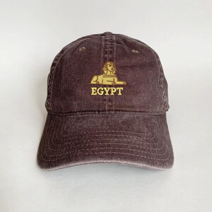 Puede incluir: Gorra de béisbol marrón con visera curva. Presenta un diseño bordado dorado de la Esfinge y la palabra "EGYPT" en oro. La gorra tiene un aspecto envejecido y vintage.
