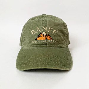 May include: A green cotton baseball cap with an embroidered logo that says "Banff National Park" with a mountain range and a sun graphic.