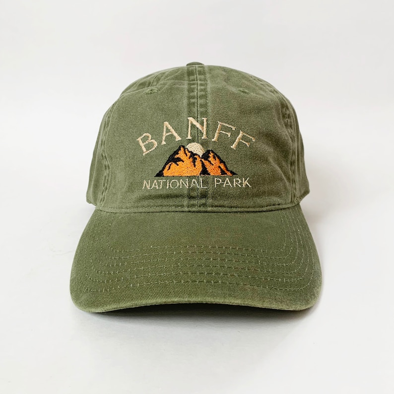 May include: A green cotton baseball cap with an embroidered logo that says "Banff National Park" with a mountain range and a sun graphic.