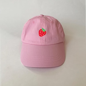 Puede incluir: Gorra de béisbol rosa con visera curva. La gorra presenta una fresa bordada con frutos rojos y hojas verdes en la parte delantera. El sombrero está hecho de una tela suave y tiene una corona redondeada.