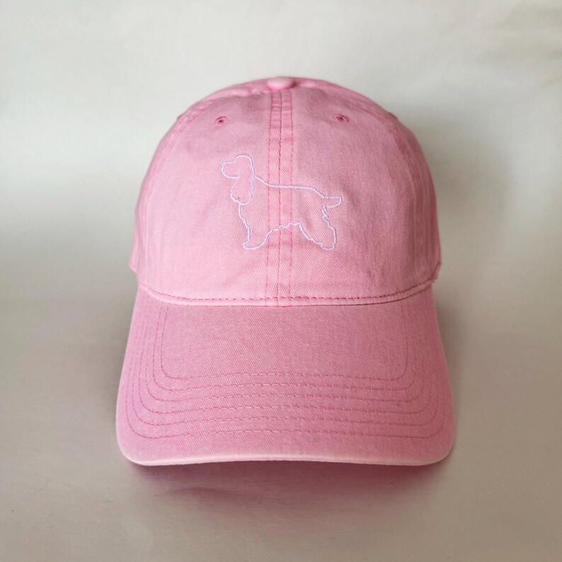 May include: A pink baseball cap with a white embroidered outline of a cocker spaniel.