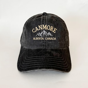 Pode incluir: Boné de beisebol preto com um design bordado branco que diz "Canmore Alberta Canada" com um gráfico de cordilheira.