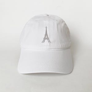 Puede incluir: Gorra de béisbol blanca con un diseño gris de la Torre Eiffel. La gorra tiene una visera curva y una correa ajustable. El diseño de la Torre Eiffel es un contorno geométrico simple.