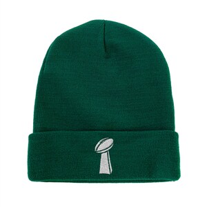 May include: A dark green beanie with a folded brim. The beanie features a white embroidered image of a football trophy on the front. The hat is made of a knitted material.