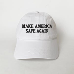 May include: White baseball cap with the text "MAKE AMERICA SAFE AGAIN" in black block letters. The cap has a curved brim and a fabric strap closure on the back. The hat is made of a soft, durable material.