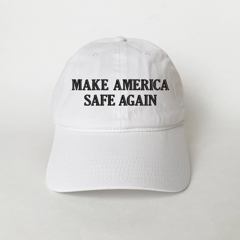 May include: White baseball cap with the text "MAKE AMERICA SAFE AGAIN" in black block letters. The cap has a curved brim and a fabric strap closure on the back. The hat is made of a soft, durable material.