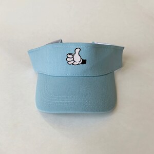 May include: A pale blue visor featuring a thumbs-up hand graphic. The visor has a curved peak and a white interior. The thumbs-up graphic is embroidered in black and white.