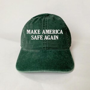 May include: A dark green baseball cap with the text "MAKE AMERICA SAFE AGAIN" in white block letters. The cap has a curved brim and a slightly faded appearance, suggesting a vintage or worn look. The hat is made of a soft fabric.