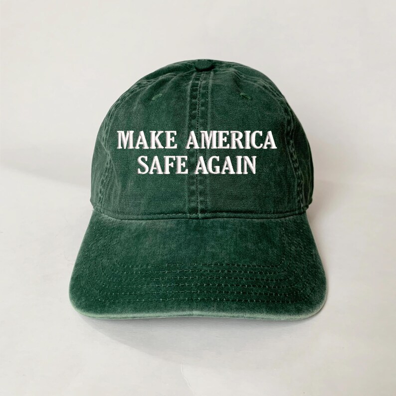 May include: A dark green baseball cap with the text "MAKE AMERICA SAFE AGAIN" in white block letters. The cap has a curved brim and a slightly faded appearance, suggesting a vintage or worn look. The hat is made of a soft fabric.